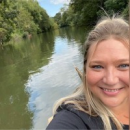 A woman smiling at the camera while on a boat or by the water, with trees and greenery reflected in the calm river behind her, capturing a peaceful Cybericon2024 moment.