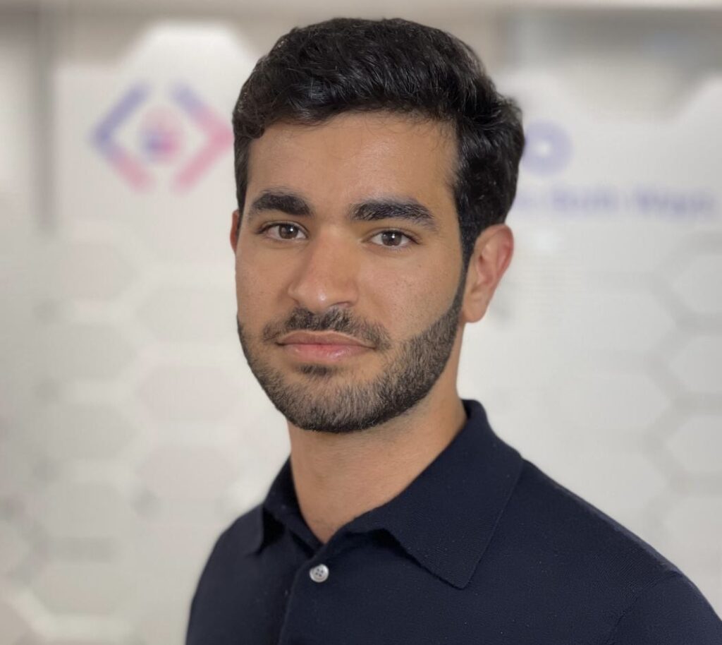 A man with short dark hair and a trimmed beard, wearing a black collared shirt, stands in front of a blurred background featuring geometric patterns and CyberWeek 2024 logos.
