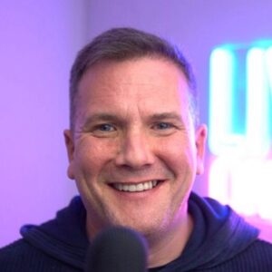 A man with short hair smiles at the camera in front of a blurred neon sign, wearing a dark hoodie, with a microphone visible in the foreground as Memcyco features on InfoSec Live's Podcast - Part 2.