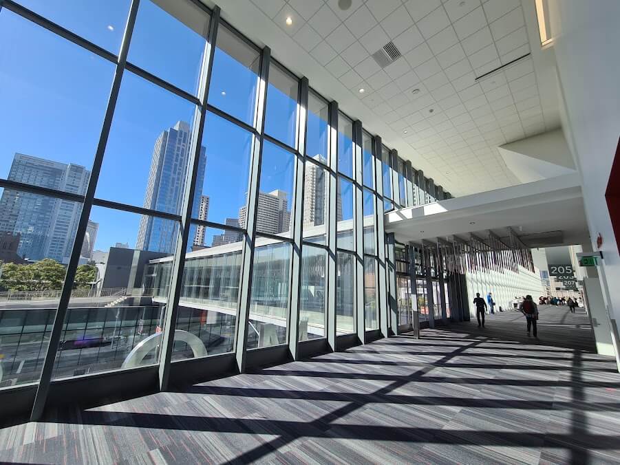 Spacious hallway with floor-to-ceiling windows showing city skyscrapers outside; a few people, including attendees of Memcyco at RSA Conference 2025, walk inside the modern building.